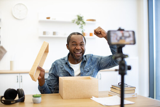 A cheerful creator records an unboxing, raising a fist beside an open cardboard package while a smartphone on a tripod sits nearby