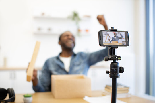 A smiling Black creator films a hands-on demonstration at a worktable, with a smartphone on a tripod, cardboard box, and wooden piece in view