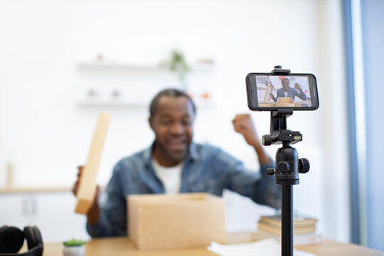 A creator records a cheerful desk presentation with a smartphone on a tripod, cardboard parcel, books, and workspace details in the background