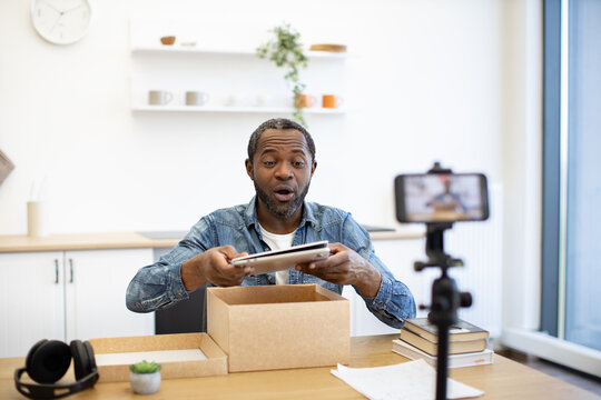 An African American man looks surprised while opening a tablet box at his desk, with a phone on a tripod recording in a bright home workspace
