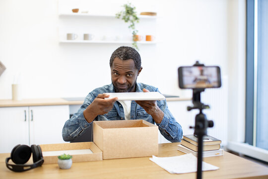 A man records an unboxing at a home desk, carefully lifting a tablet from cardboard packaging beside a smartphone mounted on a small tripod