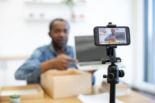 A man films himself with a smartphone on a tripod while opening a cardboard package beside a laptop in a bright home workspace at a desk