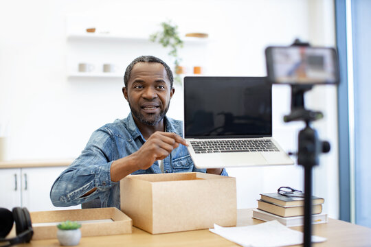 A bearded presenter gestures toward a portable computer beside open cardboard packaging, with a phone on a tripod and books on the desk in a bright room