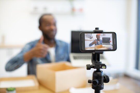 An African American creator records a product presentation at home, with a smartphone on a tripod, an open shipping box, and a laptop nearby