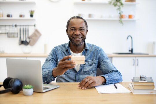 An African American man smiles at his desk while holding a small card, with a laptop, headphones, notebook, and books in a bright home workspace
