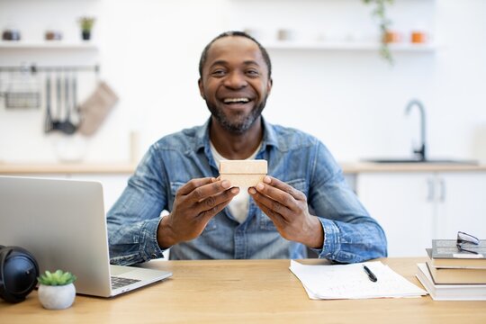 A smiling African American man sits at a desk in a bright home workspace, holding a small blank rectangular object beside a laptop and notebook