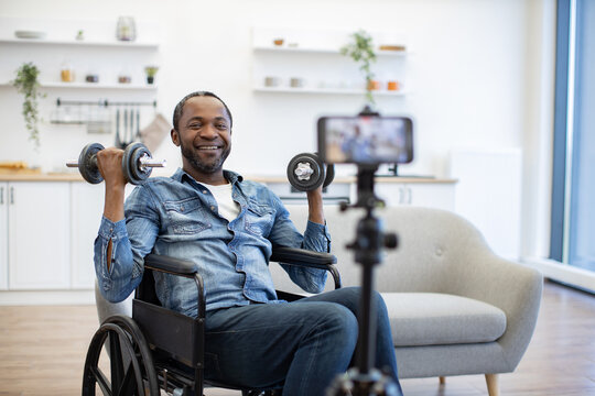 A smiling African American man exercises with dumbbells in a wheelchair while recording his strength training session at home on a smartphone