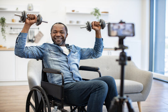 A smiling man in a wheelchair lifts dumbbells at home while a smartphone on a tripod records his seated strength workout in a bright living room