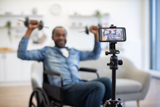 A man in a wheelchair lifts dumbbells while recording a home workout on a smartphone tripod in a bright, modern living room with natural daylight