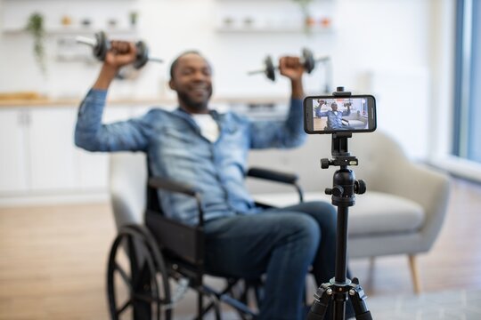 A man in a wheelchair exercises with hand weights while a smartphone on a tripod records his workout in a bright, comfortable living space