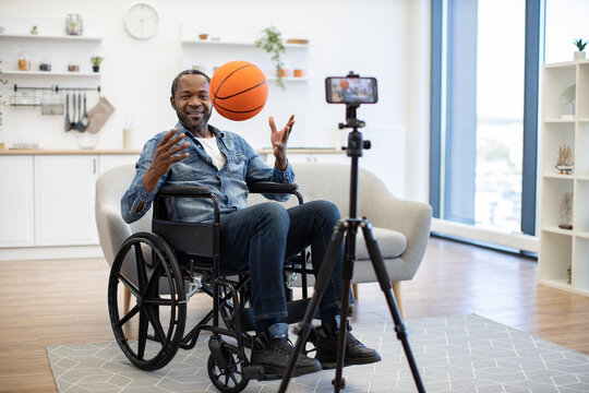 An African American wheelchair user tosses a basketball while filming himself at home, showcasing adaptive recreation and video creation