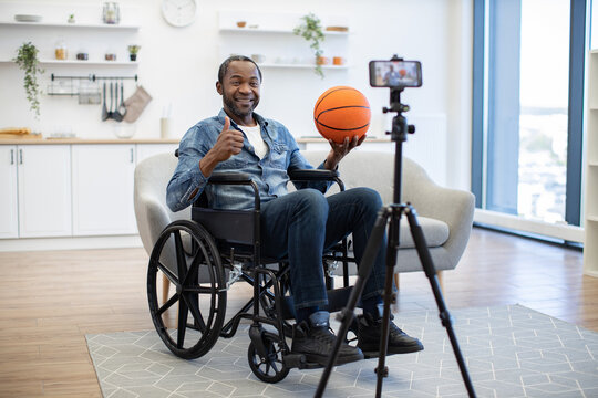 A smiling wheelchair user holds a basketball and gives a thumbs-up while filming at home, highlighting accessible recreation and inclusive digital content