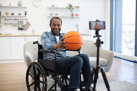 A smiling man in a wheelchair holds an orange basketball while a smartphone on a tripod records him in a bright modern living space indoors