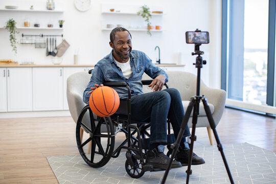 A smiling man in a wheelchair holds a basketball while recording a video at home, with a smartphone mounted on a tripod in the bright living room