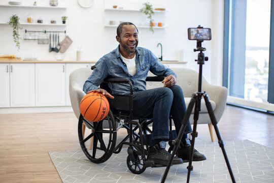 A smiling man records a home video beside a basketball, seated in a wheelchair in a bright living room with a smartphone mounted on a tripod