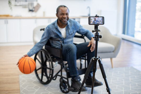 A smiling man records himself at home beside a basketball, using a smartphone mounted on a tripod while seated in a wheelchair in a bright living room