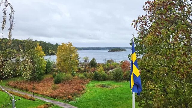 Beautiful view of Swedish flag waving beside autumn trees and wooded islands against cloudy sky. Sweden.