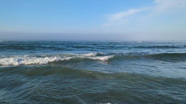 Sea waves roll and splash white foam and water droplets under blue hazy sky