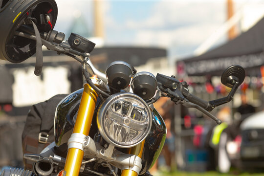 Close-up of motorcycle front showing round headlight, gold front forks, handlebars, and mounted helmet strap at outdoor event.