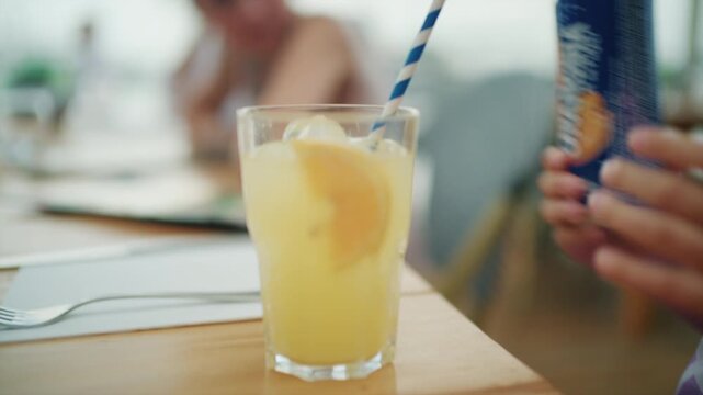Close-up of a glass of orange soda with an orange slice and striped paper straw as a girl in a star T-shirt touches it, French Riviera, France.