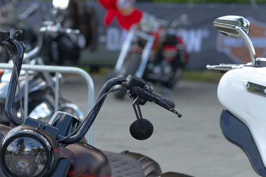 Close-up of motorcycle handlebar with mirror, throttle control, and headlight in outdoor parked setting with other motorcycles blurred in background.