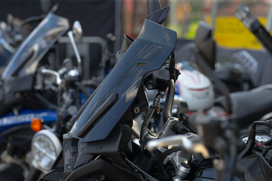 Close-up view of motorcycle handlebar area showing windshield, control cables, key switch, and instrument cluster with other parked motorcycles blurred in background.