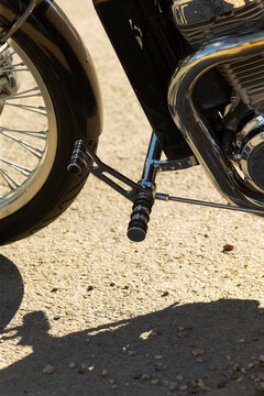 Close-up of motorcycle footpeg, front wheel and chrome exhaust on gravel surface with visible shadow and sunlight reflections.