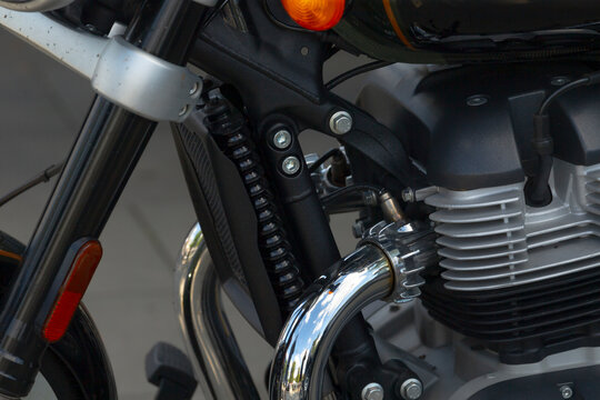 Close-up of motorcycle engine and front fork showing chrome exhaust pipe, cooling fins, mounting bolts, and shock spring detail.