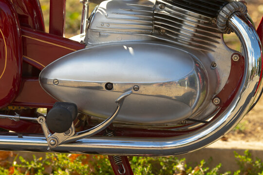 Close-up of motorcycle engine and chrome exhaust showing metal casing, foot pedal, and part of red frame in natural light.