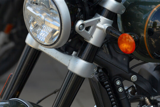 Close-up of motorcycle front forks, headlight, and orange turn signal showing metal clamps, black suspension tubes, and wiring.