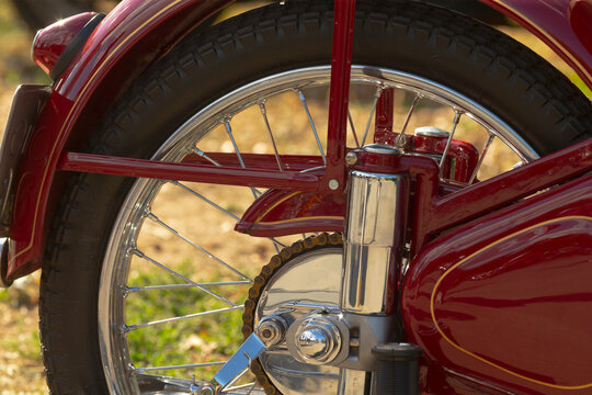Close-up of rear wheel, chain and suspension on red vintage motorcycle showing chrome hub, spokes and painted fender in daylight.