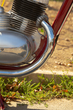 Close-up of motorcycle engine showing chrome exhaust pipe, finned cylinder head, and red painted frame on outdoor ground with small plants.