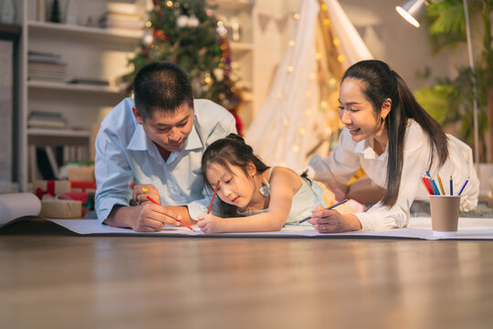 Asian parents and little daughter drawing with colored pencils on floor at Christmas at home
