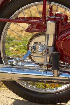 Close-up of rear wheel, chain, and chrome exhaust of red motorcycle showing spokes and suspension mounting hardware on sunny pavement.