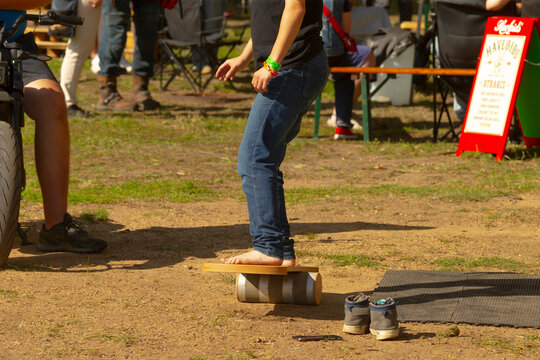 Person balancing barefoot on wooden board placed over cylindrical roller on dirt ground at outdoor event with shoes and mat nearby.