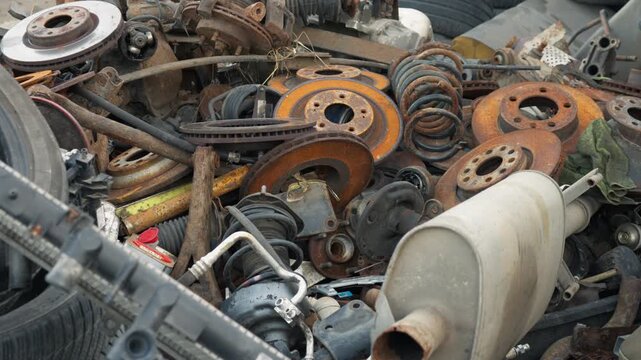 Discarded vehicle scrap metal and old car tires accumulate in a junkyard heap. Weathered exhaust muffler and rusted iron parts await sorting at waste disposal facility.