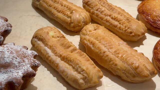 Various types of fresh sweet pastries and eclairs are displayed on parchment paper counter at bakery. Traditional french dessert, homemade baking goods, delicious sugar treats, coffee shop display.