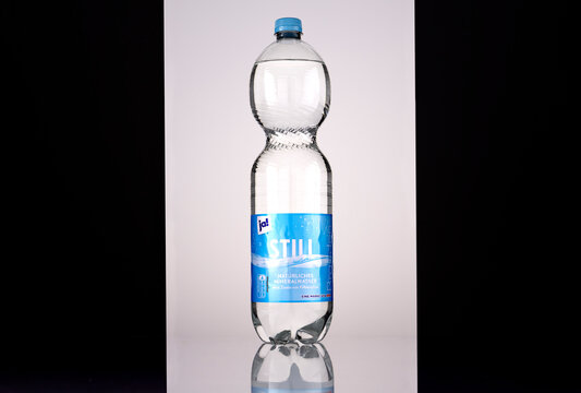 Front view of a ja! still natural mineral water bottle with a blue screw cap and blue German-language label identifying it as a REWE brand. Backlit studio photograph on a reflective surface against a 