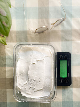 Homemade Greek yogurt in a glass container on a digital scale with eyeglasses on a plaid tablecloth. Perfect for healthy breakfast and clean eating concepts.
