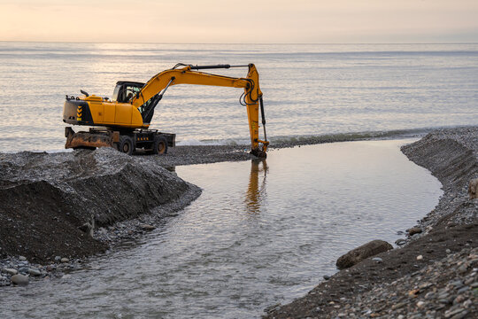 Excavator clears and deepens coastal river mouth after heavy rains carried mud, gravel and stones downstream. Dredging restores water flow and removes sediment accumulated where river enters sea.