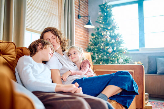 Loving mother kissing her son's head while daughter cuddles next to her on a sofa in a cozy living room during holidays