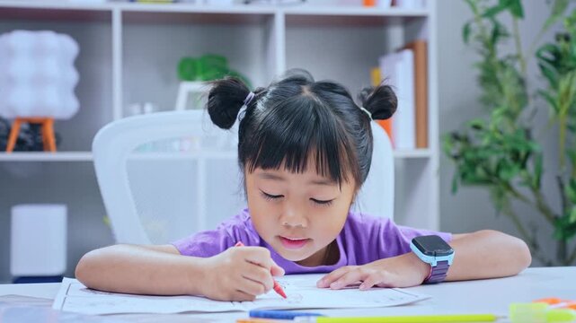 Asian girl student writing homework attentively with red pen at desk in living room for primary education concept