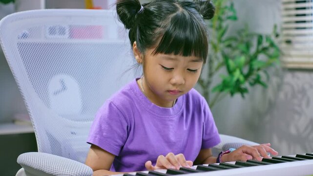 Asian girl playing synthesizer keyboard with focused attention during music lesson at home