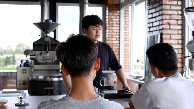 shot of a friendly barista explaining specialty coffee to two customers at a roastery cafe counter with a large roasting machine in the background