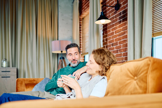 Middle-aged couple sitting on a comfortable sofa in their modern living room, talking and enjoying hot coffee together