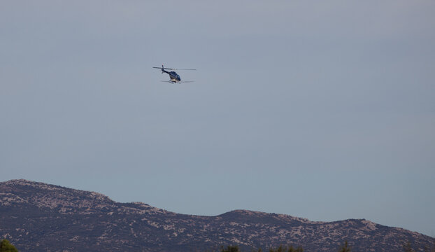 Un h&eacute;licopt&egrave;re vole dans un ciel clair au-dessus d'une r&eacute;gion montagneuse ou collinaire, avec une v&eacute;g&eacute;tation visible au premier plan.