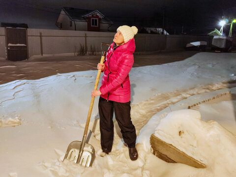 Woman shoveling snow in a residential yard at night. Winter storm cleanup and snow removal. Cold weather home maintenance. Seasonal outdoor work and safety