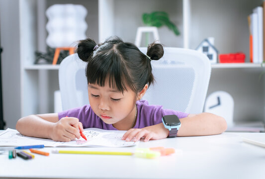 Asian girl focused on writing homework and drawing with colored pencil on table at home