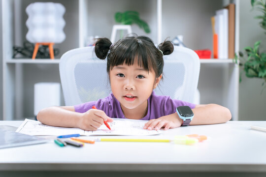 Asian girl focused on writing homework and drawing with colored pencil on table at home