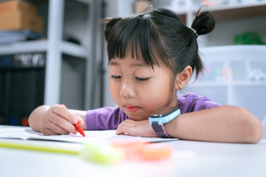 Asian girl focused on writing homework and drawing with colored pencil on table at home